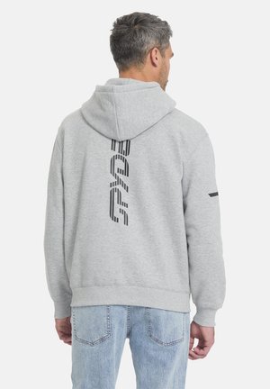 Mann trägt hellgrauen Kapuzenpullover mit vertikalem schwarzem Grafiktext auf dem Rücken, kombiniert mit hellblauen Jeans, mit dem Rücken zum Betrachter.