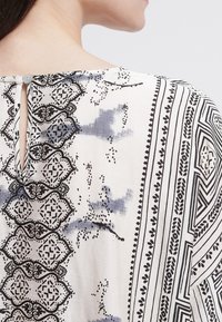 Flödande, vit tygblus med intrikata svarta och grå blommönster samt geometriska mönster, med en nyckelhålrygg och knappstängning.