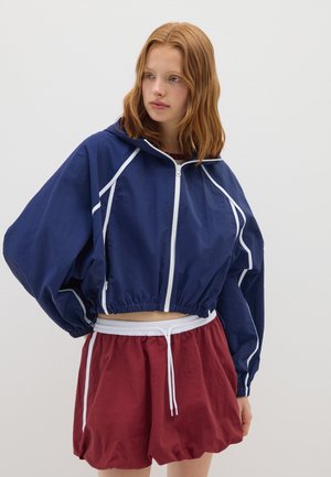 Joven mujer con cabello rojo, vistiendo una sudadera cortada con cremallera color azul marino y pantalones cortos flojos color burdeos con cordones blancos, en un fondo sencillo.
