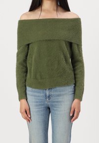 Maglione color verde oliva scollato sulle spalle, realizzato in materiale morbido e peloso, con maniche lunghe e orlo inferiore aderente, abbinato a jeans azzurri chiari.