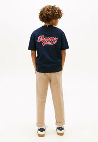 Marineblå bomulds t-shirt med et stort rødt "Tommy" logo og teksten "Class of 1985" på bagsiden, parret med beige bukser og sneakers.