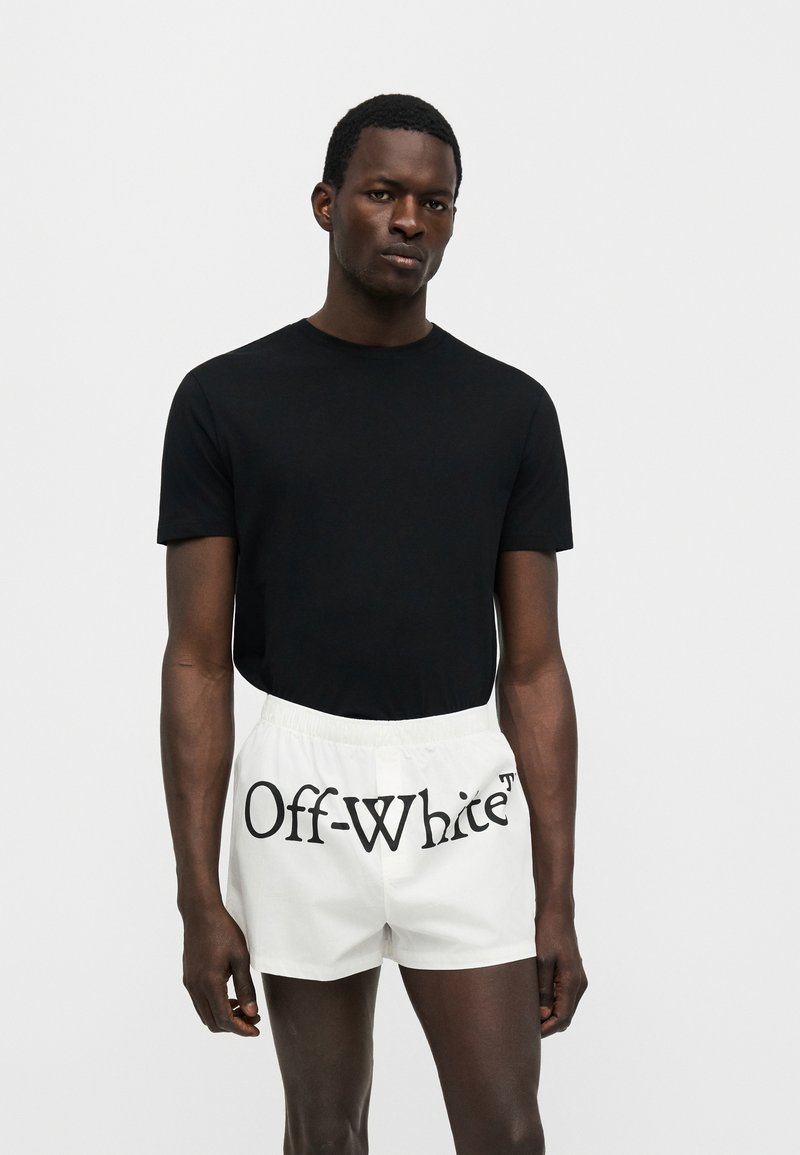 Muž v čiernom tričku s krátkym rukávom a bielych krátkych nohaviciach s potlačeným logom „Off-White“ vpredu na jednoduchom pozadí.