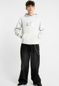 Jasnoszara bluza z kapturem z metalicznym grafiką z przodu, zestawiona z luźnymi czarnymi spodniami z detalem w postaci łańcucha.