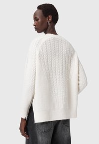 Donna che indossa un maglione bianco a maglia texturizzata con un motivo intrecciato sulla schiena, abbinato a jeans scuri su uno sfondo neutro.