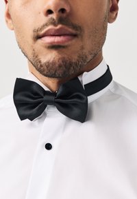 Papillon in raso nero su una camicia bianca, caratterizzato da un design classico con una texture liscia e un nodo prominente al centro.