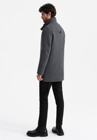 Abrigo de lana gris con un cuello alto, detalle de yugo en la parte posterior, combinado con pantalones negros y elegantes botas negras. Textura suave con un ajuste a medida.