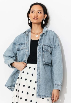 Femme portant une veste en jean bleu clair oversize, un haut noir, une jupe blanche à pois noirs, des boucles d'oreilles blanches en forme de goutte et un collier doré avec pendentif fleur.