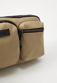 Borsa in nylon beige con finitura texturizzata, dotata di un compartimento principale e di una tasca più piccola con zip. Cerniere e dettagli neri inclusi.