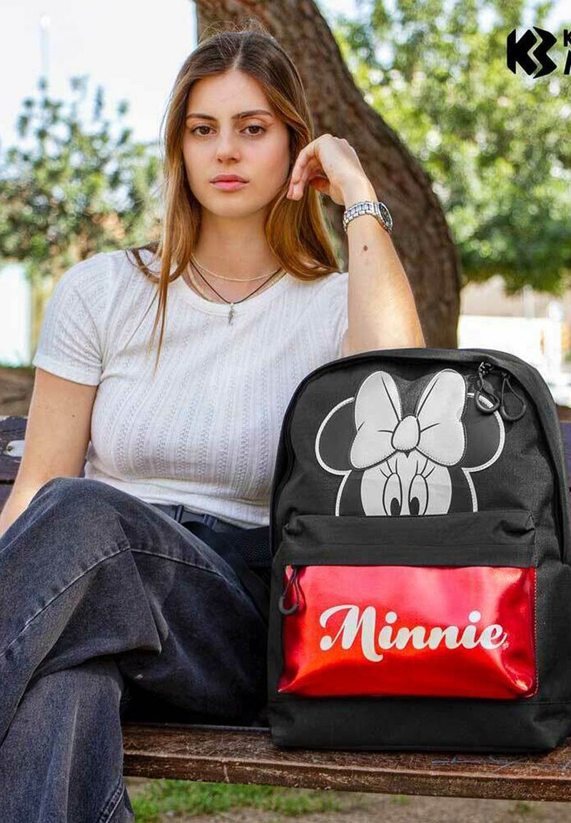 Ung kvinde iført hvid skjorte og mørke jeans sidder på en bænk i parken ved siden af en sort rygsæk med Minnie Mouse-ansigt og en rød "Minnie" pung.