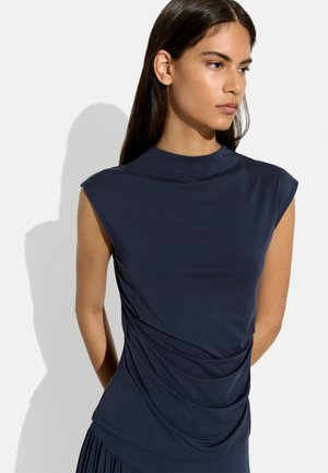 Młoda kobieta z długimi ciemnymi włosami, ubrana w granatowy top bez rękawów z marszczonymi detalami, stojąca na jasnym tle.