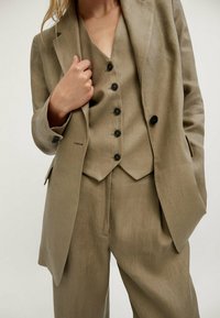 Beige liinapuku, johon kuuluu muotoiltu bleiseri, napitteellinen liivi ja leveät housut, joissa on sileä tekstuuri ja hienovarainen kiilto.