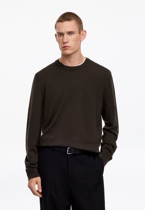 Junger Mann mit kurzen Haaren, der einen dunkelbraunen Langarmpullover und schwarze Hosen vor einem einfarbigen weißen Hintergrund trägt.