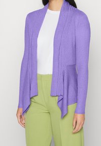 Cardigan côtelé lavande à l'avant ouvert, ourlet allongé et manches ajustées, porté avec un haut blanc et un pantalon vert.