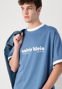 Blue cotton t-shirt with white crew neckline and sleeve trims. Front features printed text "calvin klein" and "New York City." Denim jacket draped over arm.