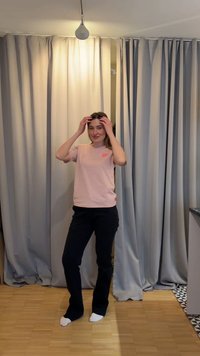 Νέα γυναίκα που διορθώνει τα μαλλιά της, στέκεται ξυπόλητη σε ξύλινο πάτωμα μπροστά από κλειστές μπεζ κουρτίνες με φωτισμό από πάνω.