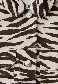 Close-up af beige stof med sorte zebra-striber, metaltrykknapper indgraveret med "Street One" og påsyede lommepatches.