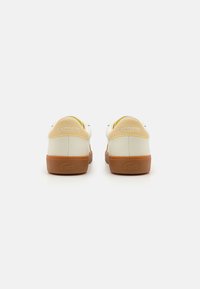 Fehér bőr sportcipő, gumi talppal, a sarokban feltűnő Lacoste logóval, lekerekített formával és minimalista design részletekkel.