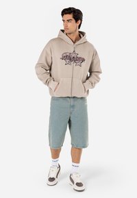 Beige zip-up hoodie with graphic print, paired with light blue denim shorts. Model wears white and gray sneakers, with white socks featuring logo.