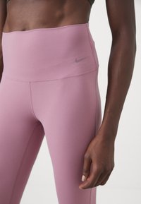Femme portant un legging lilas taille haute avec un petit logo Nike sur la hanche gauche, debout avec le bras droit détendu le long du corps.