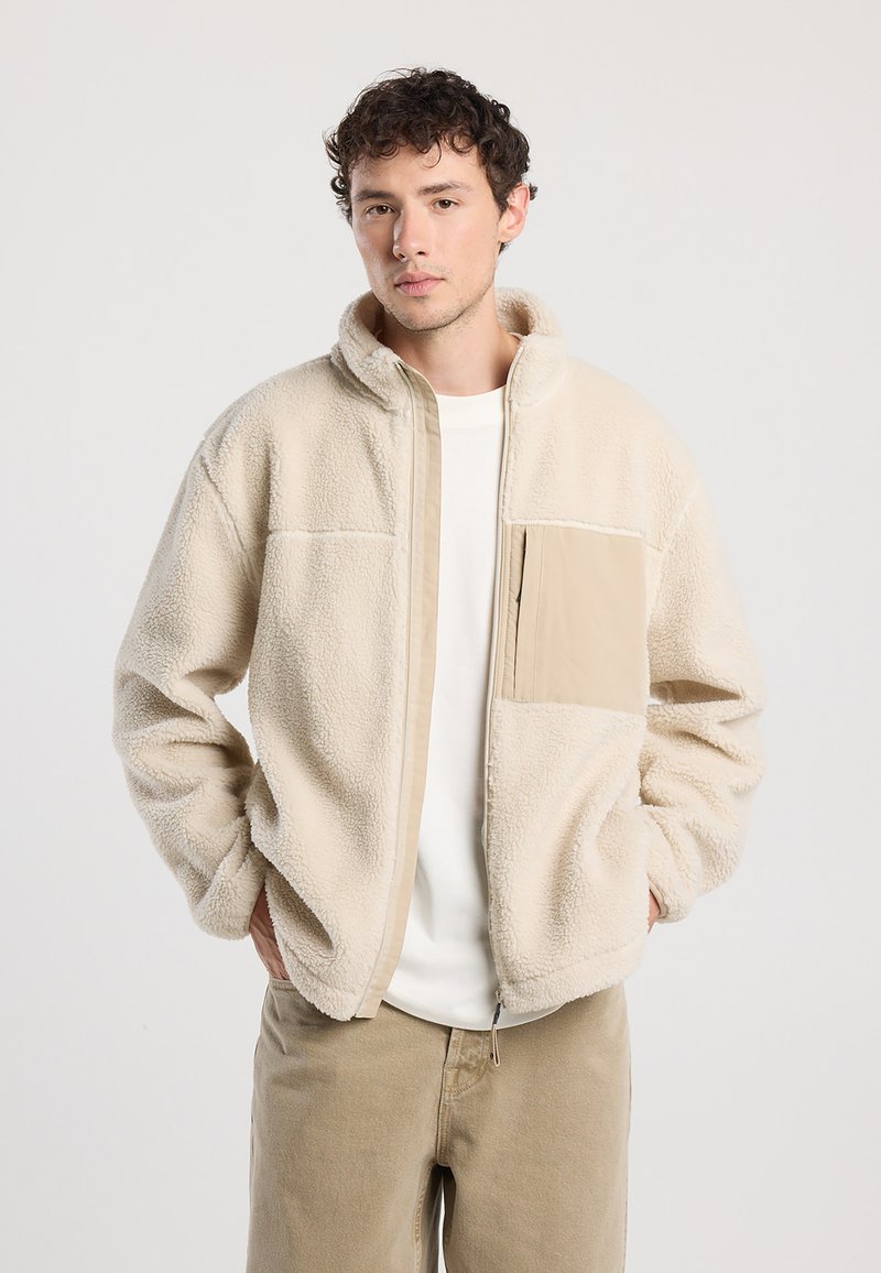 Beige Fleecejacke mit Reißverschluss vorne, hohem Kragen und einer hellbraunen Tasche. Sie hat eine weiche Textur und einen lässigen Schnitt und wird über einem weißen Hemd getragen.