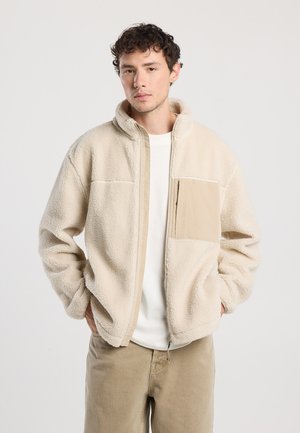 Beige fleece jas met een rits aan de voorkant, hoge kraag en een tan kleurige zak. Heeft een zachte textuur en een ontspannen pasvorm, gedragen over een wit shirt.