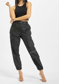 Zwarte cargobroek met meerdere zakken, ritsdetails, elastische boorden en een relaxte pasvorm, gecombineerd met een strak zwart cropped top en doorzichtige hakken.