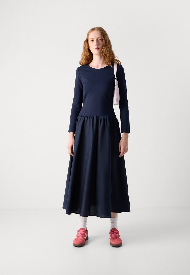 Robe longue à manches longues de couleur marine avec taille froncée et jupe fluide. Associée à des baskets roses et un petit sac rose pâle. Texture de tissu lisse.