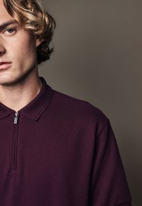 Polo shirt bordeaux avec une finition texturée, manches courtes et un col zippé. Le tissu semble doux et léger.
