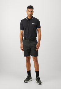 Μαύρο polo μπλουζάκι με λευκές λεπτομέρειες, λογότυπο στο στήθος, συνδυασμένο με μαύρες βερμούδες. Μοντέλο που φοράει μαύρες κάλτσες και γκρι αθλητικά παπούτσια.