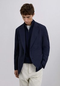 Blazer azul marino con un corte entallado, diseño de un solo botón, cuello de solapa, combinado con un suéter oscuro y pantalones de color claro. Textura de tela suave.