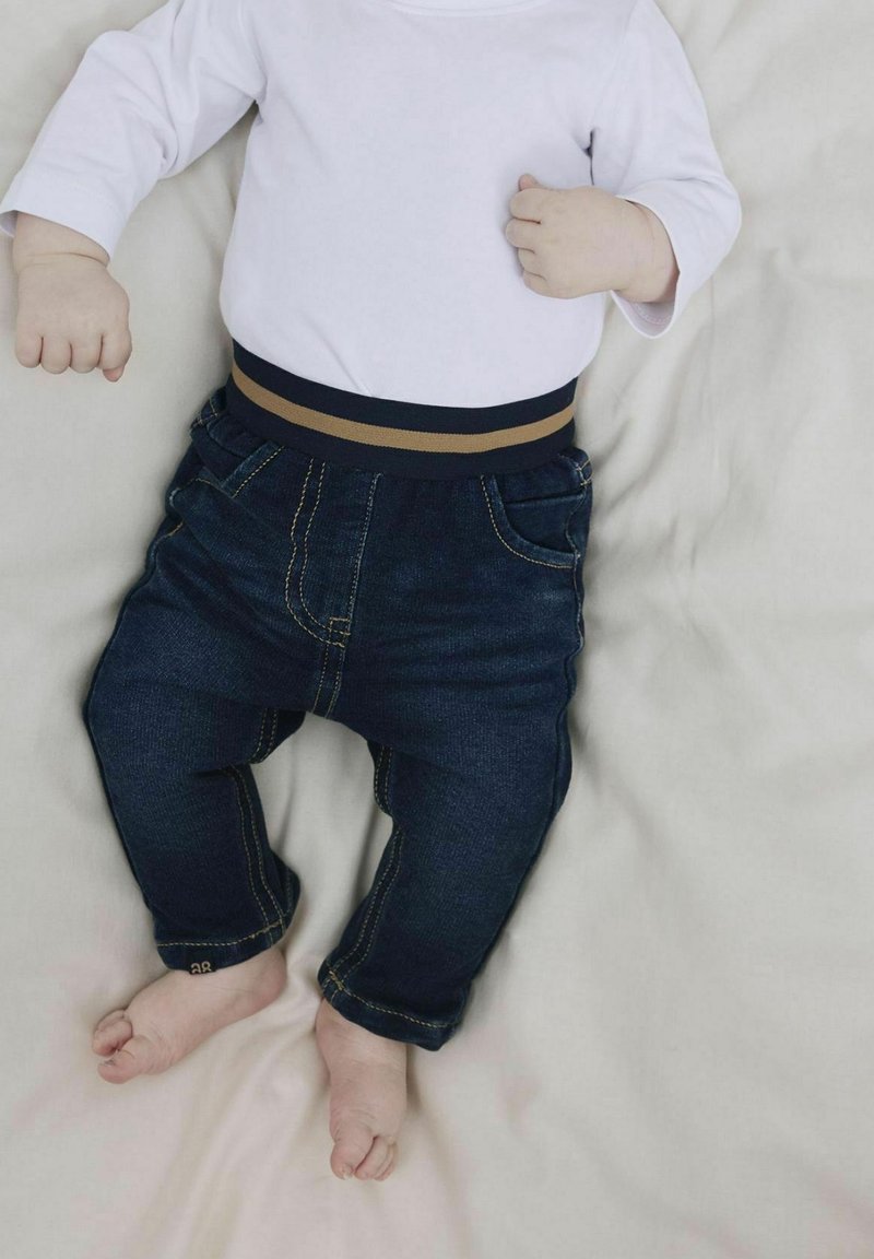Bébé allongé sur une surface claire, portant une chemise blanche à manches longues et un pantalon en jean bleu foncé, pieds nus visibles.