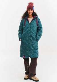 Dlouhý petrolejový prošívaný kabát s diamantovým prošíváním, vínovými detaily, předním zipem a růžovou nášivkou s logem, nošený přes hnědé kalhoty a boty.