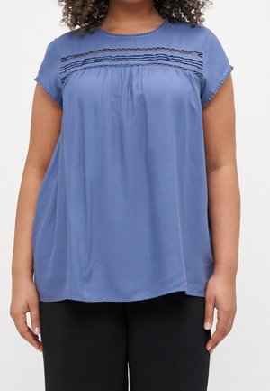 Femme portant un chemisier bleu ample à manches courtes avec un détail en dentelle sur la poitrine et un pantalon noir, debout devant un fond uni.
