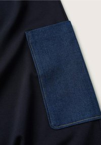 Tela azul marino con un bolsillo rectangular de mezclilla; el bolsillo tiene costuras en contraste y una textura suave, que complementan el fondo más oscuro.