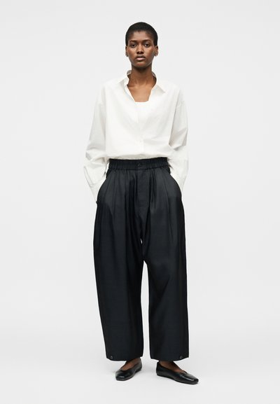 Γυναίκα που στέκεται με τα χέρια στις τσέπες, φορώντας άνετο λευκό πουκάμισο με κουμπιά, φαρδιά μαύρη παντελόνα και μαύρα επίπεδα παπούτσια, σε λευκό φόντο.