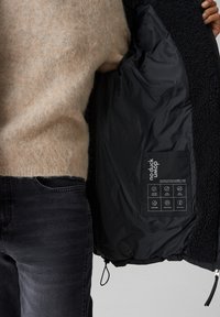 Giacca nera con un interno morbido e peloso. Presenta un'etichetta con istruzioni per la cura e icone. Il tessuto esterno appare liscio e resistente all'acqua.