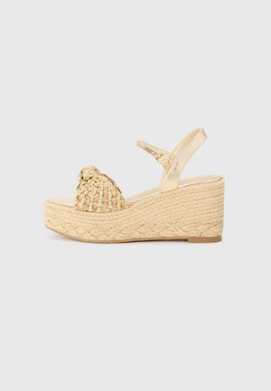 Sandales plateformes avec un upper en fibre naturelle tissée et une bride métallique dorée à la cheville. Talon en coin texturé de couleur beige clair.