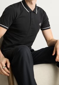 Černé polo tričko vyrobené z bavlny, s klasickým límcem s bílým lemem, krátkými rukávy a malým logem CK na levé hrudi.