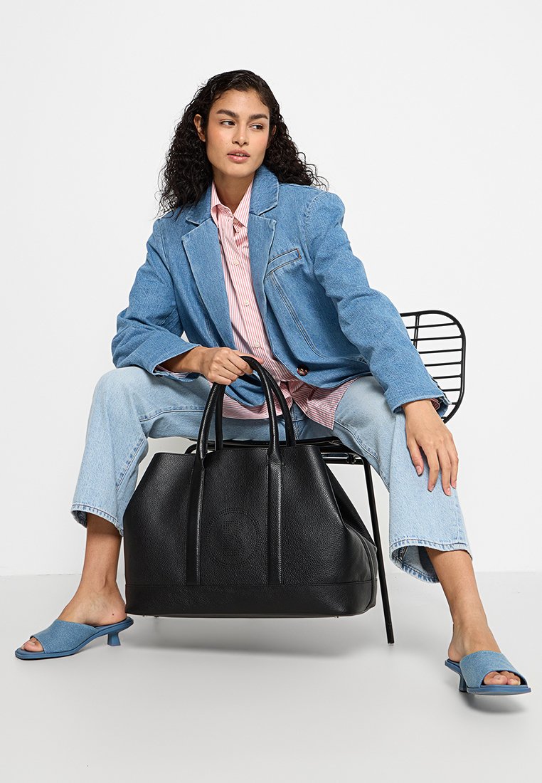 Schwarze Leder-Tote mit strukturiertem Design, doppelten Griffen, texturierter Oberfläche und minimalen Akzenten, platziert neben einer Person in legerer Kleidung.