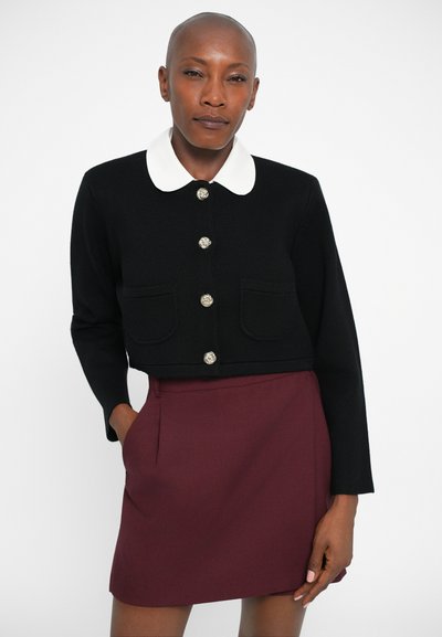 Veste noire courte avec un col blanc et des boutons argentés, associée à une jupe bordeaux. La veste possède deux poches avant.