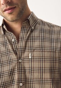 Man wearing brown and beige checkered button-up shirt with a collar and a small logo tag on the chest pocket.