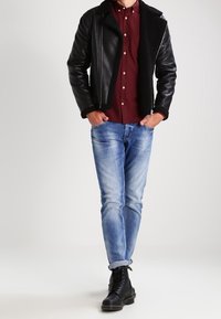 Giubbotto di pelle nera con colletto in shearling, camicia rossa borgogna con bottoni, jeans azzurri chiari e stivali neri con lacci e punta rotonda.