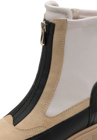 Bottine beige et noire avec fermeture à glissière, textures lisses et en daim, et couture détaillée sur le côté et la semelle.