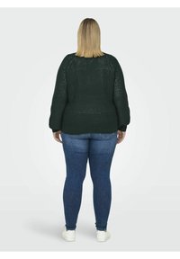 Personne aux cheveux blonds mi-longs portant un pull en maille vert foncé, un jean bleu et des baskets blanches, debout, de dos.