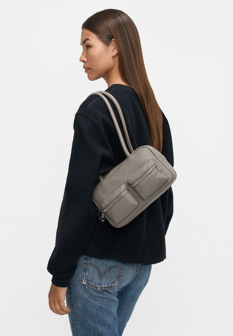 Mała szara skórzana torba na ramię z dwoma przednimi kieszeniami na zamek, regulowanymi paskami i gładkim wykończeniem, noszona na czarnym swetrze.