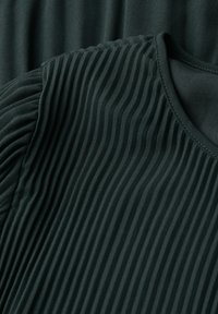 Dunkelgrüner, texturiert Stoff mit vertikalen Falten, glattem Ausschnitt und sanften Drapierungen. Nahaufnahme der Kleidungsstückkonstruktion.