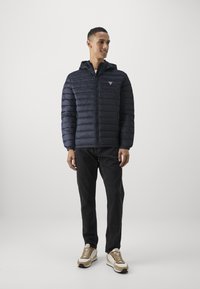 Navy puffer jacket with horizontal quilting, zip closure, and hood; paired with dark jeans and neutral-toned sneakers.