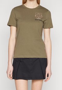 Camiseta de manga corta verde olivo con un gráfico de tigre y el texto "ALMA SALVAJE" en strass, combinada con una falda negra con pliegues.