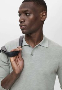 Light gray polo shirt with a textured fabric, two buttons, and a collar. A dark leather strap rests on the shoulder, held by a hand.