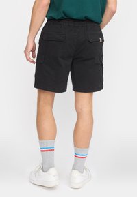 Homme portant un short cargo noir, des chaussettes grises avec des rayures rouge et bleu, et des baskets blanches, vu de dos à partir de la taille.